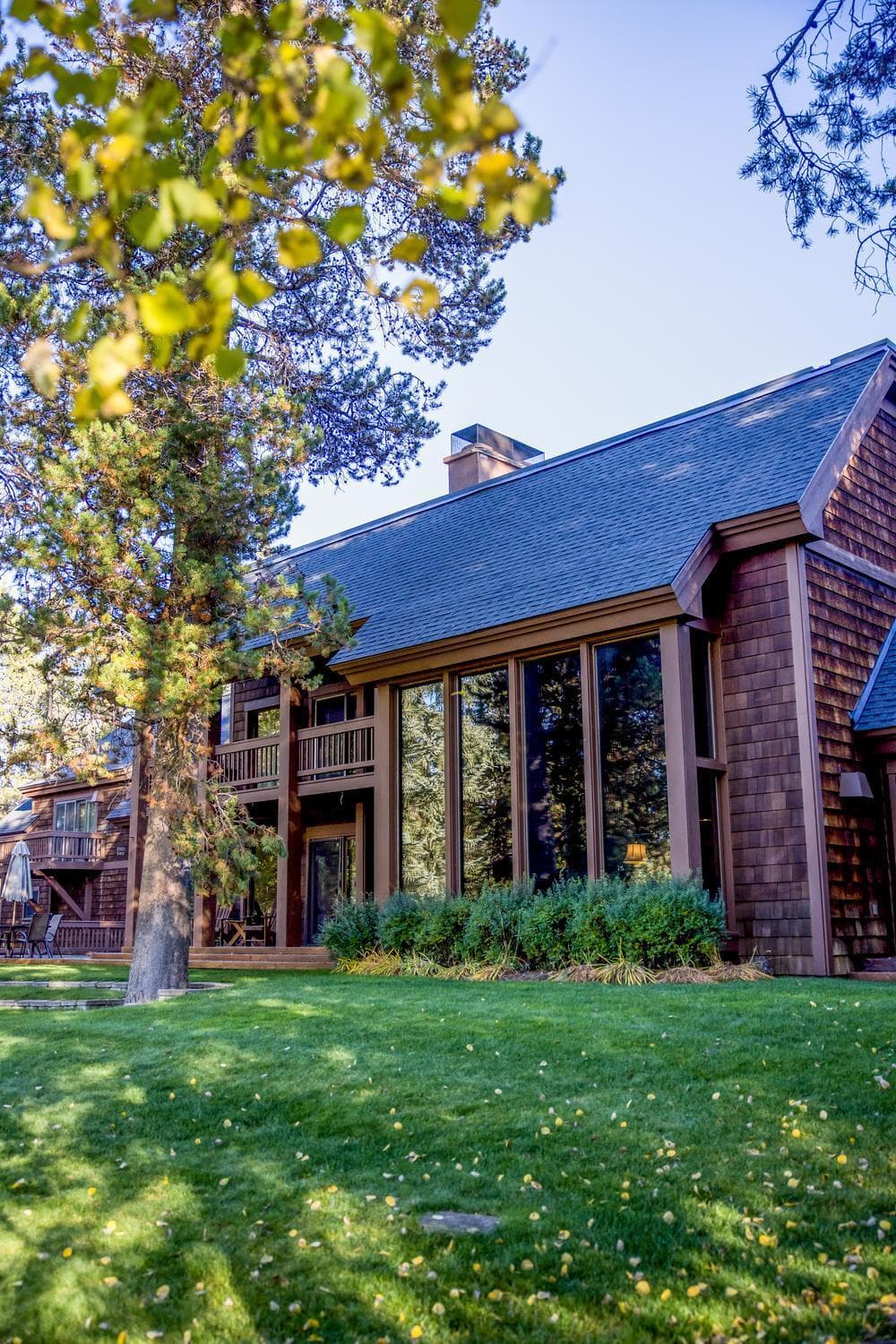 A two-story wooden house with large floor-to-ceiling windows, surrounded by green grass, trees, and shrubs, under a clear blue sky.
