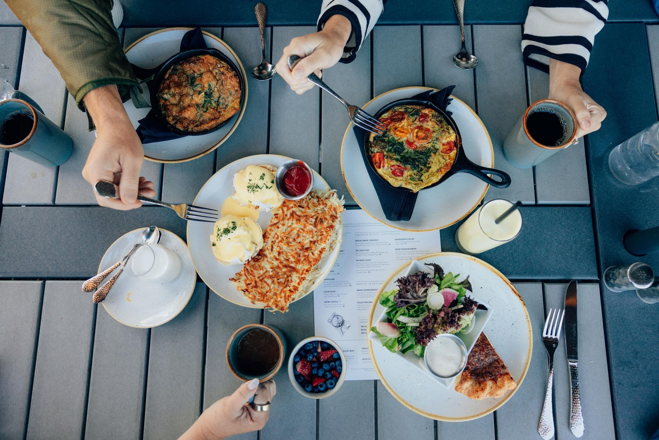 Four people share a brunch at an outdoor table with omelets, eggs benedict, hash browns, salad, pie, fruit, coffee, and water. Hands reach in with utensils, and a menu sits in the center.
