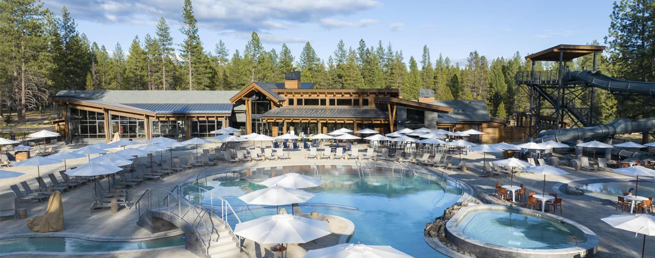 Outdoor swimming pool area with lounge chairs and umbrellas, surrounded by pine trees and a large wooden lodge. Water slides and hot tubs are visible, and the sky is partly cloudy. The scene appears calm and inviting.