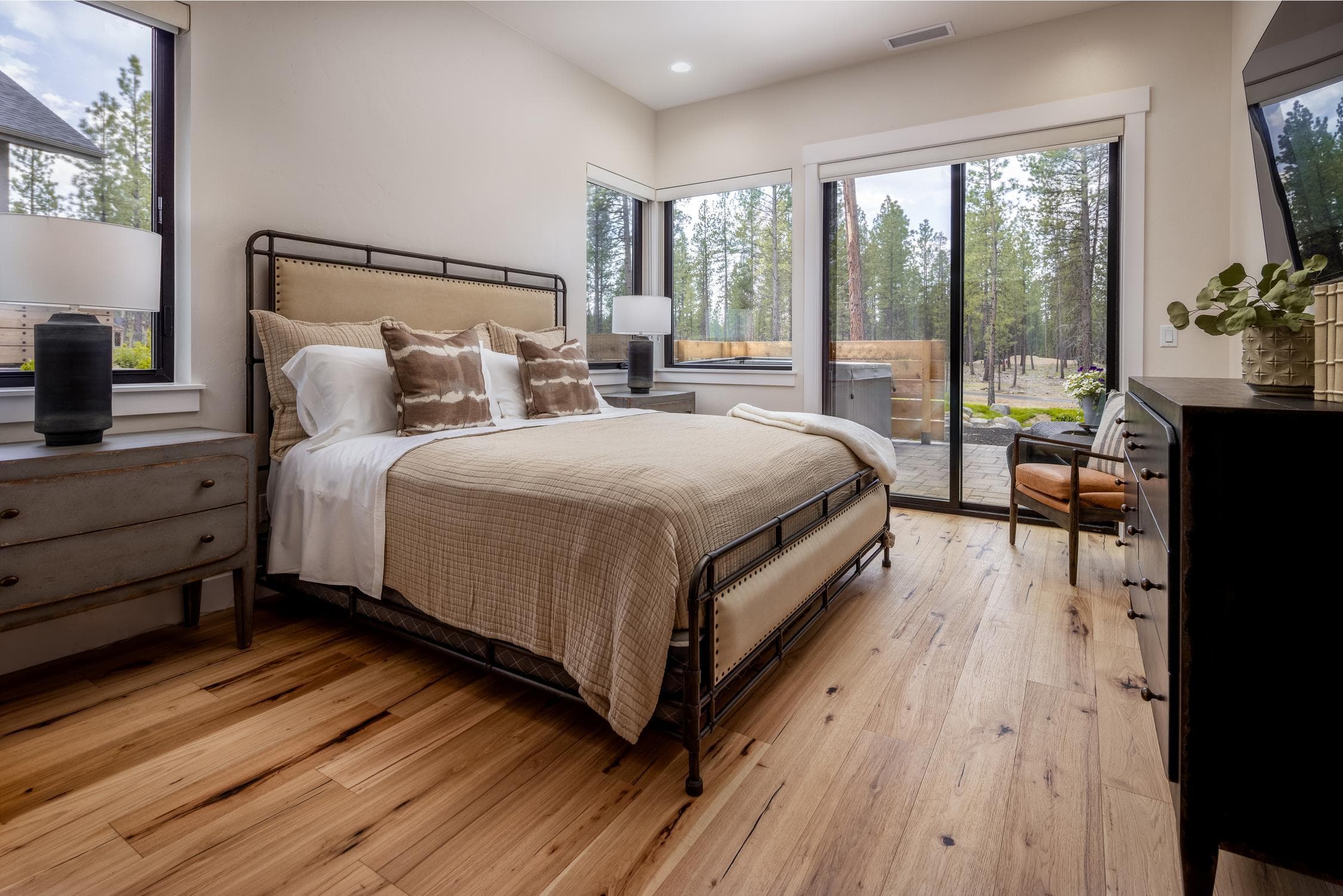A cozy bedroom with a metal bed, tan bedding, two side tables with lamps, large windows, a desk, and sliding glass doors opening to a patio and wooded view in a Forestbrook Home.