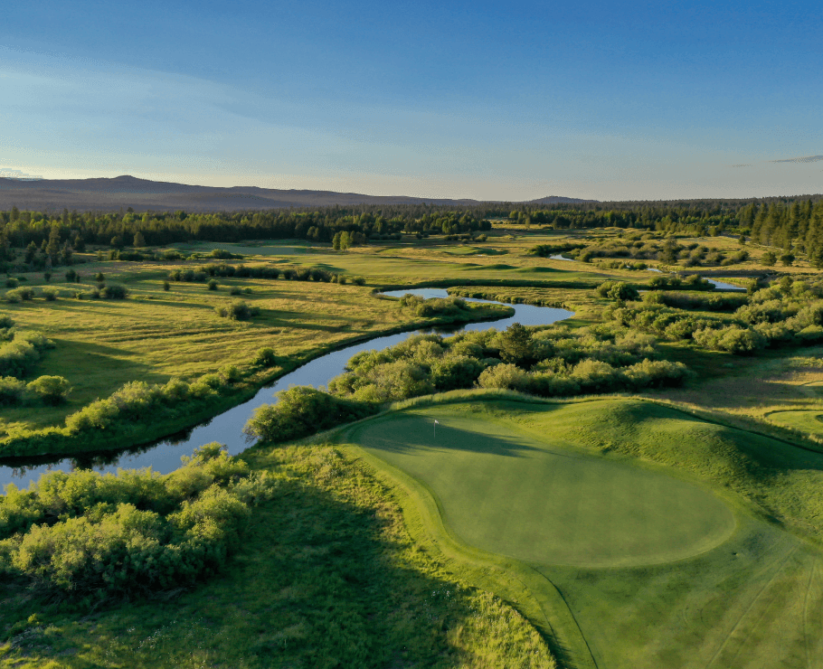 An aerial view of a lush green golf course bordered by a winding river and patches of trees, under a clear blue sky with mountains in the distance.