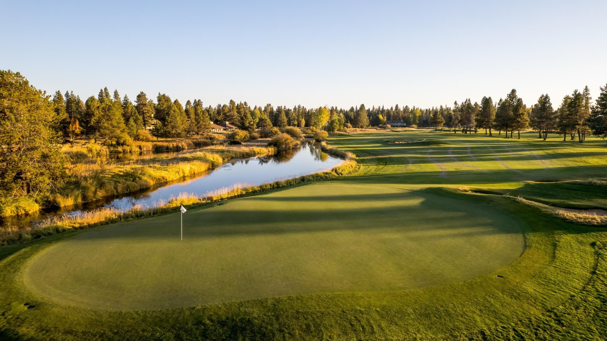 A golf course with a green, a flagstick, and a calm river running alongside, surrounded by trees and golden grass under a clear sky at sunset.