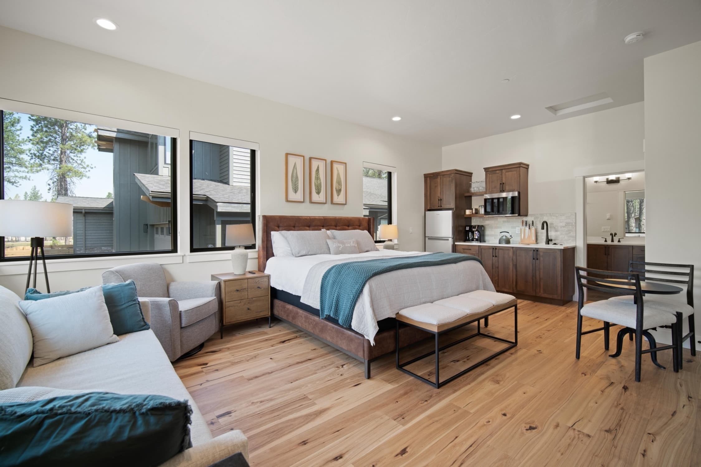 A Forestbrook studio with a bed, sofa, armchair, small kitchenette, and dining table. Large windows provide natural light. Light wood floor and neutral decor create a cozy, inviting atmosphere.
