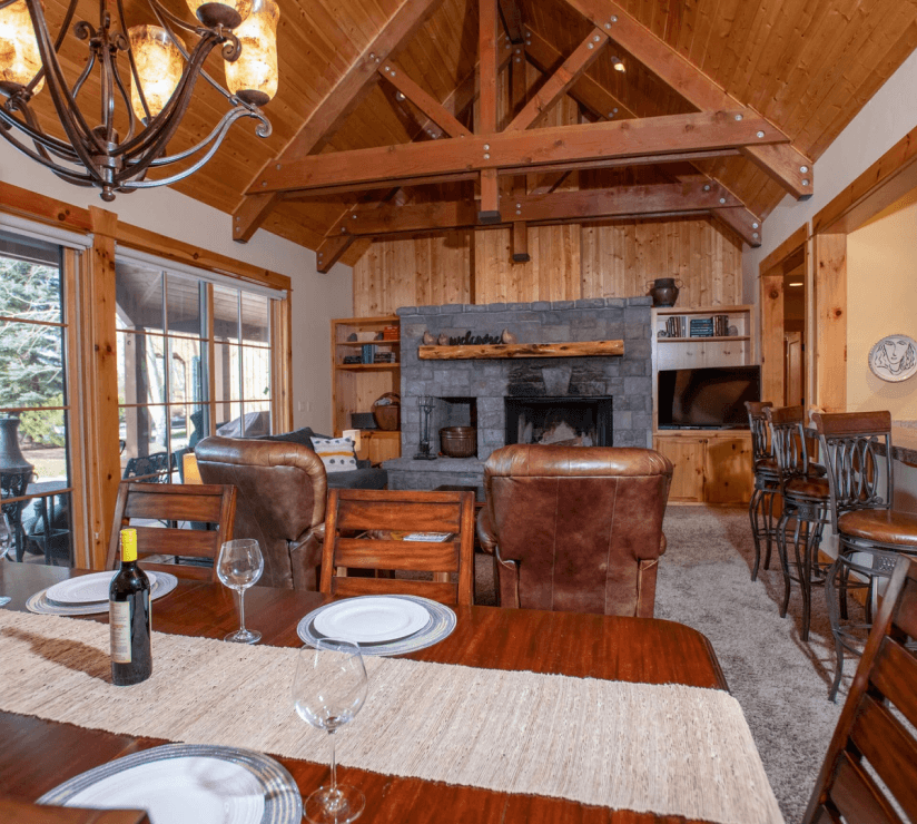 A cozy wood cabin living room with exposed beams, stone fireplace, leather chairs, a dining table set with plates and wine, large windows, bar stools, and warm lighting throughout.