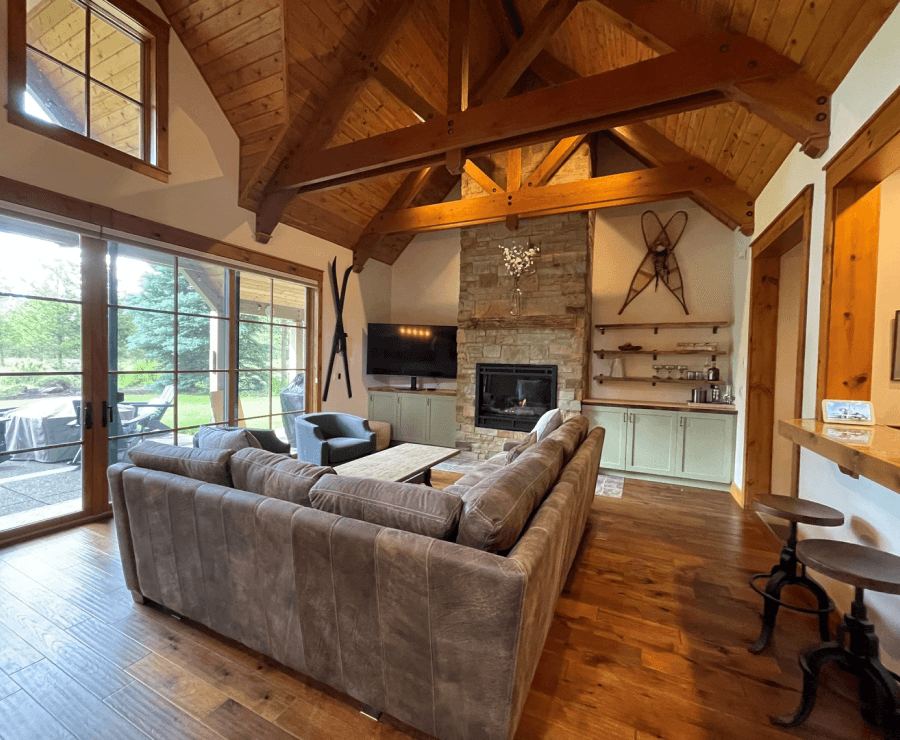Cozy living room with vaulted wood ceiling, stone fireplace, large gray sectional sofa, TV, and big windows overlooking a patio. Rustic decor includes skis on the wall and exposed beams.