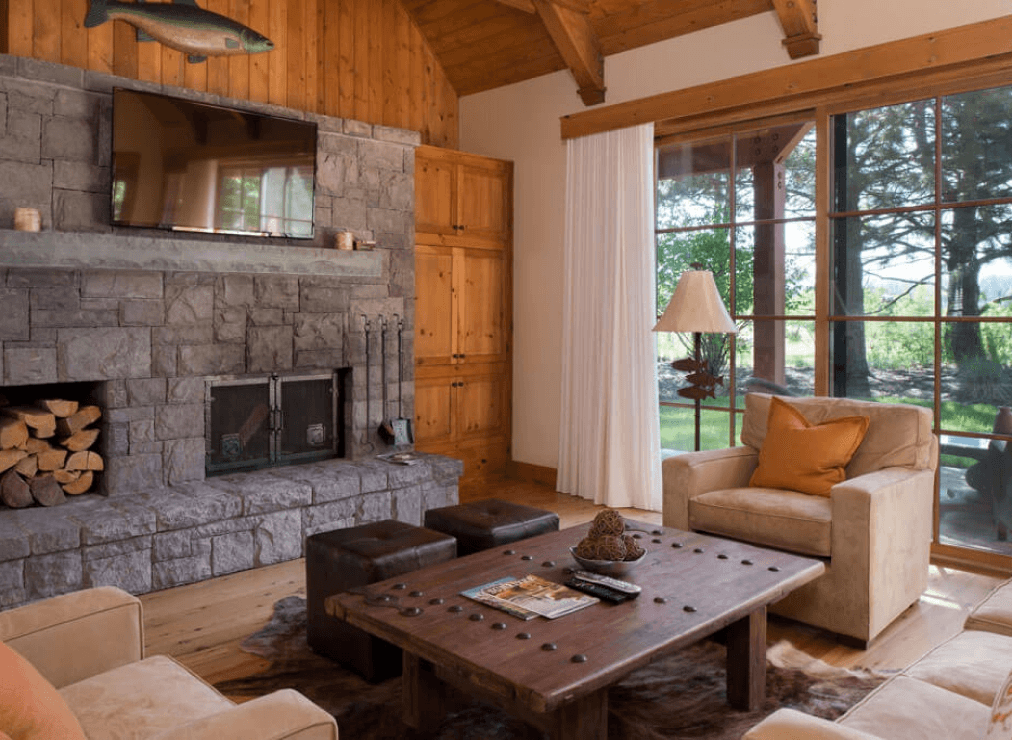 Rustic living room with stone fireplace, wood walls, mounted TV, beige armchairs, a wooden coffee table, and large windows showing a green outdoor view. Logs are stacked beside the fireplace.