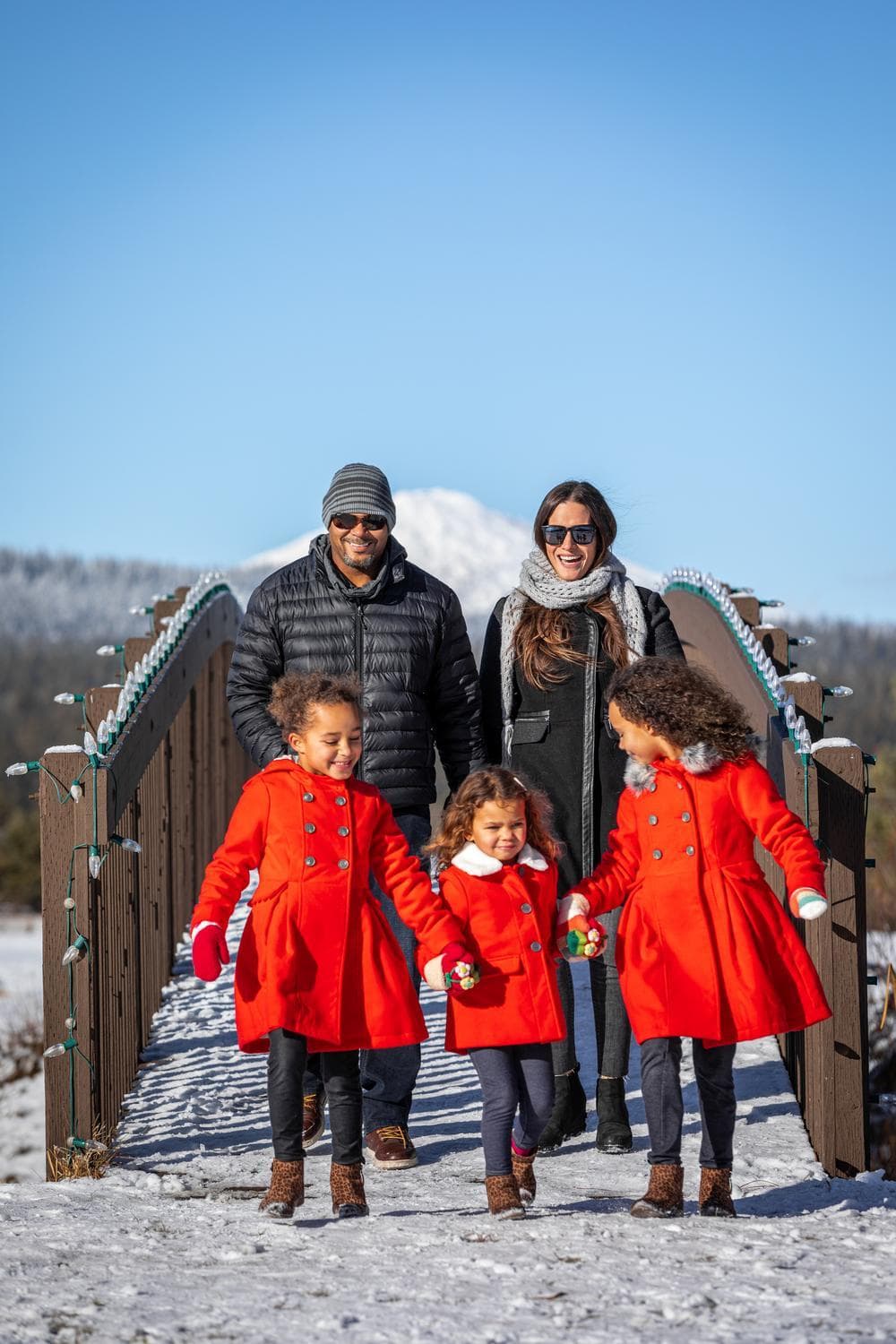 A family walks on a snowy bridge. Two adults in winter clothes smile behind three young girls in matching red coats, holding hands. A mountain and blue sky are in the background.