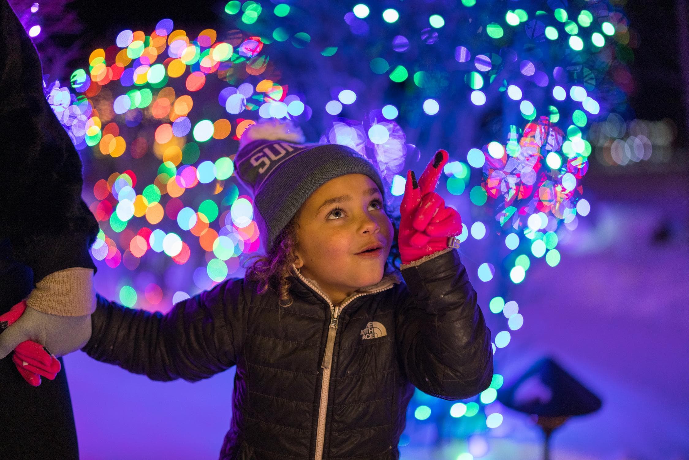 A young child in winter clothes excitedly points at colorful holiday lights in the background, holding an adults hand. The scene is bright and festive.