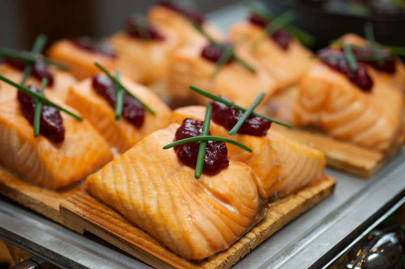 Several pieces of cooked salmon are served on wooden planks, each topped with a dollop of red sauce and garnished with two crisscrossed chive stems.