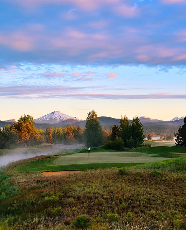 A golf course green with a flag, surrounded by trees and mist, with snow-capped mountains in the background under a colorful sunrise sky.