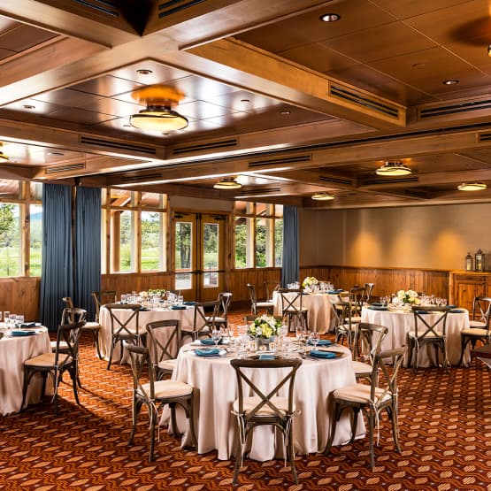Elegant banquet hall with round tables set for an event, each table decorated with floral centerpieces and place settings. Large windows, wood paneling, and patterned carpet create a warm, inviting atmosphere.