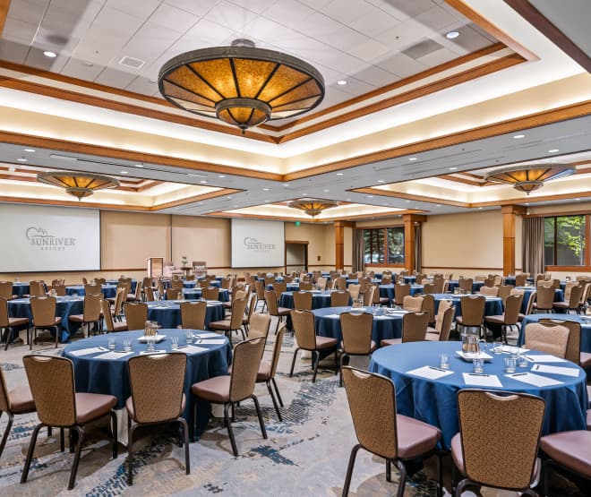 Spacious, well-lit conference room with round tables covered in blue cloths, surrounded by chairs. Projector screens display Sunriver Resort. Large ceiling lights and windows provide ample lighting.