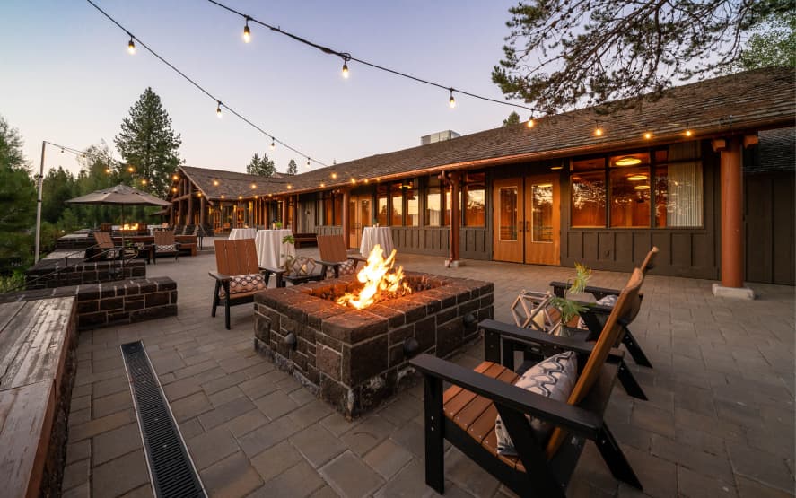 Outdoor patio at sunset with string lights, wooden chairs around a stone fire pit, tables with white cloths, and a rustic building in the background surrounded by trees.