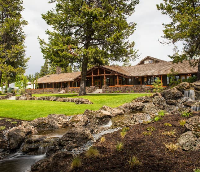 A rustic lodge with a stone chimney sits behind a lush lawn and landscaped garden, featuring rocks, trees, and a small stream with a waterfall in the foreground.