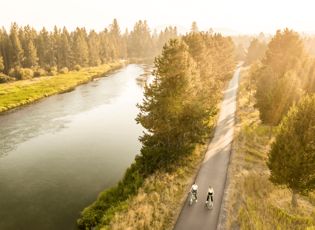 Two people ride bicycles on a paved path beside a river, surrounded by tall trees and grass, under warm, golden sunlight.