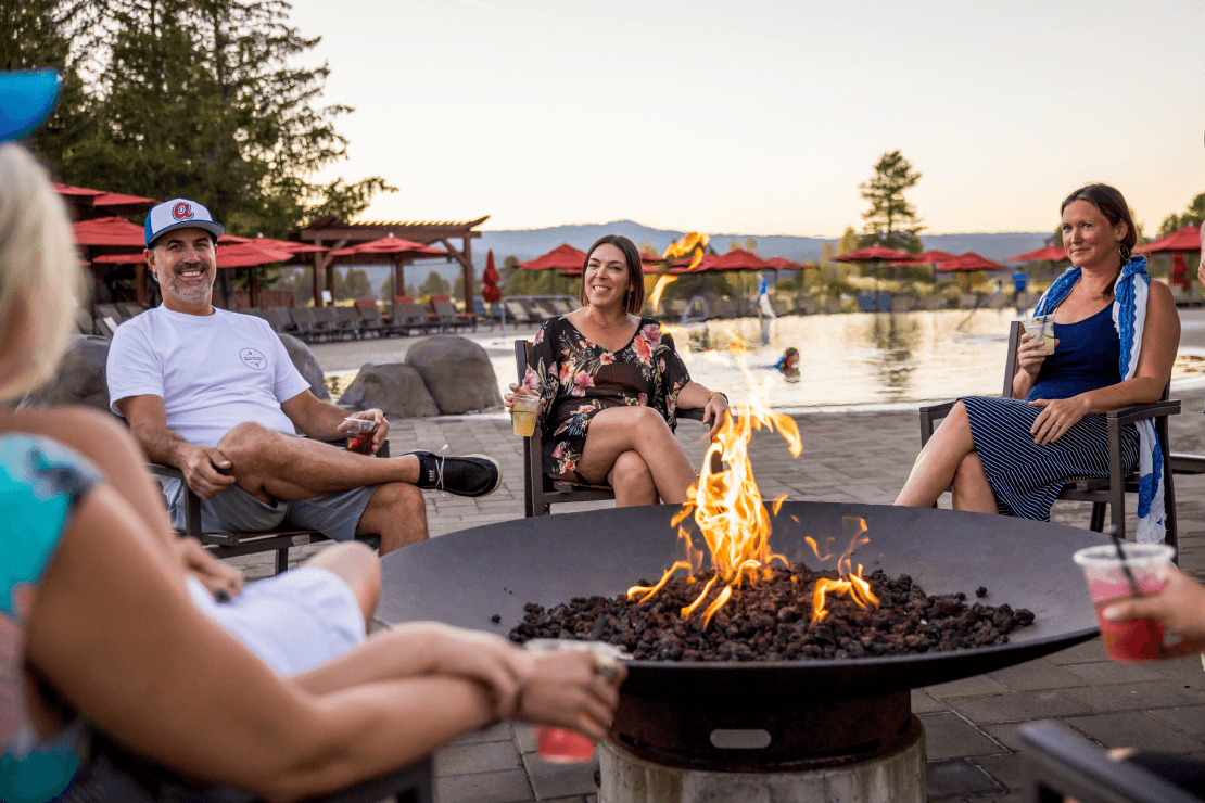 Four people relax around a fire pit, smiling and holding drinks, near a pool with red umbrellas and trees in the background at sunset.