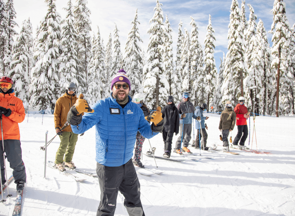 A smiling person in a blue jacket and purple beanie poses with gloved hands up in front of a group of people on skis, surrounded by snowy trees on a bright winter day.
