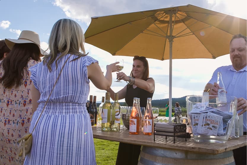 People gather outdoors at a wine stand with bottled drinks and wine glasses on a barrel table under a large umbrella. A woman serves wine while others chat. The sky is partly cloudy.