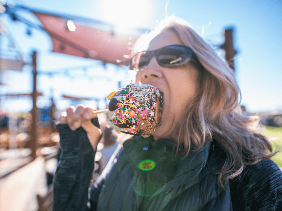 A woman wearing sunglasses eats a large chocolate-covered treat with colorful sprinkles outdoors on a sunny day.