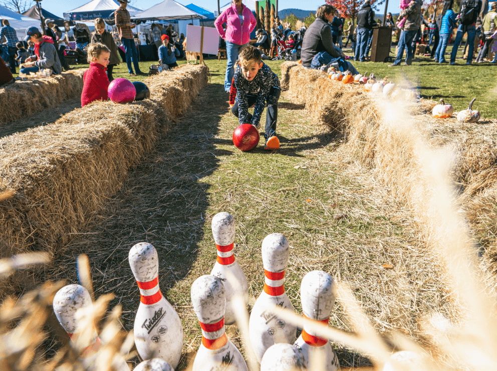 A child rolls a red bowling ball toward white pins set up on grass between hay bales, with pumpkins nearby. Other children and adults watch at an outdoor fall festival under sunny skies.