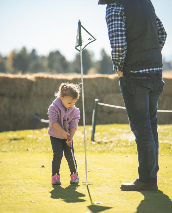 A young girl in a pink jacket putts a golf ball on a green as an adult stands nearby, watching her.