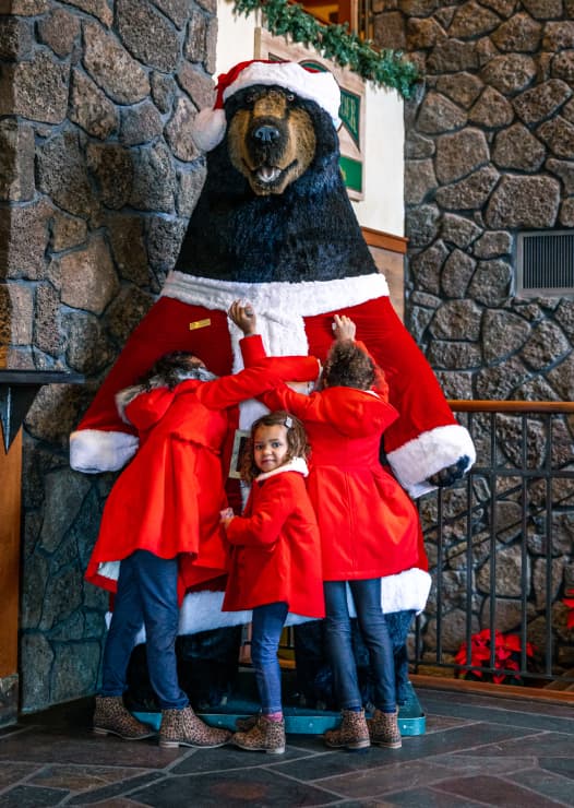 Three children in red coats hug a large bear statue dressed in a Santa suit, indoors by a stone wall decorated with greenery and holiday accents.