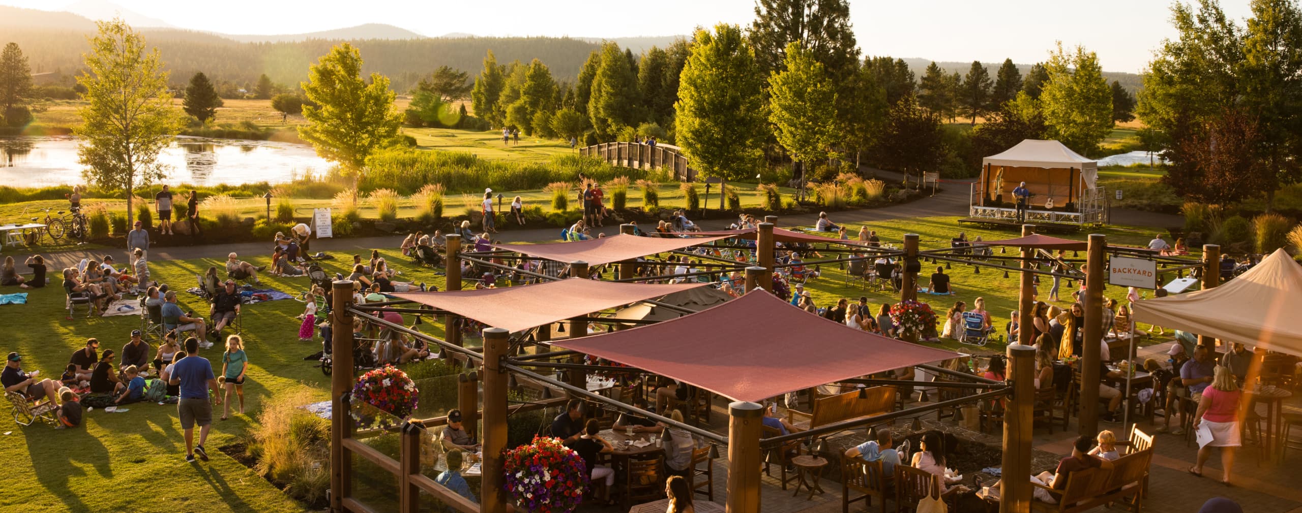 People gather on a grassy lawn near a river at sunset, enjoying an outdoor concert with a band on stage. Families sit on blankets and chairs, surrounded by trees and patio structures.