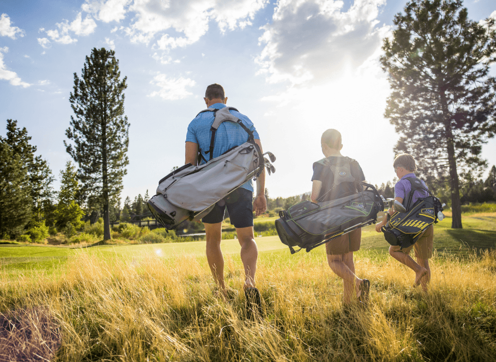 Three people carrying golf bags walk through tall grass toward a sunlit golf course, with trees and a bright sky in the background.