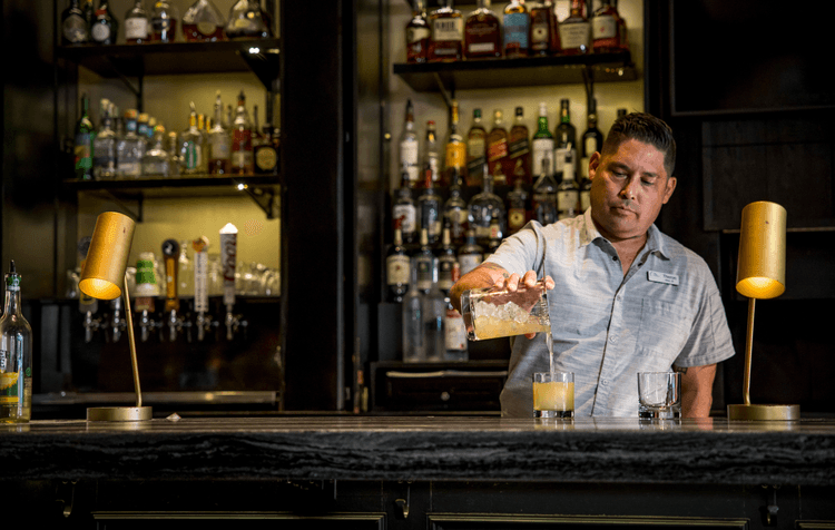 A bartender pours a cocktail from a mixing glass into a tumbler at a dimly lit bar, with shelves of liquor bottles and two golden lamps on the counter.