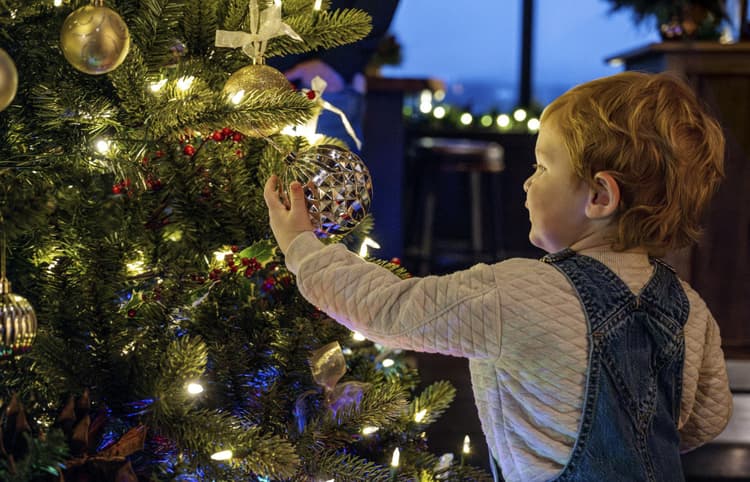 A young child in overalls reaches to hang a shiny ornament on a lit Christmas tree adorned with decorations, glowing with warm lights.