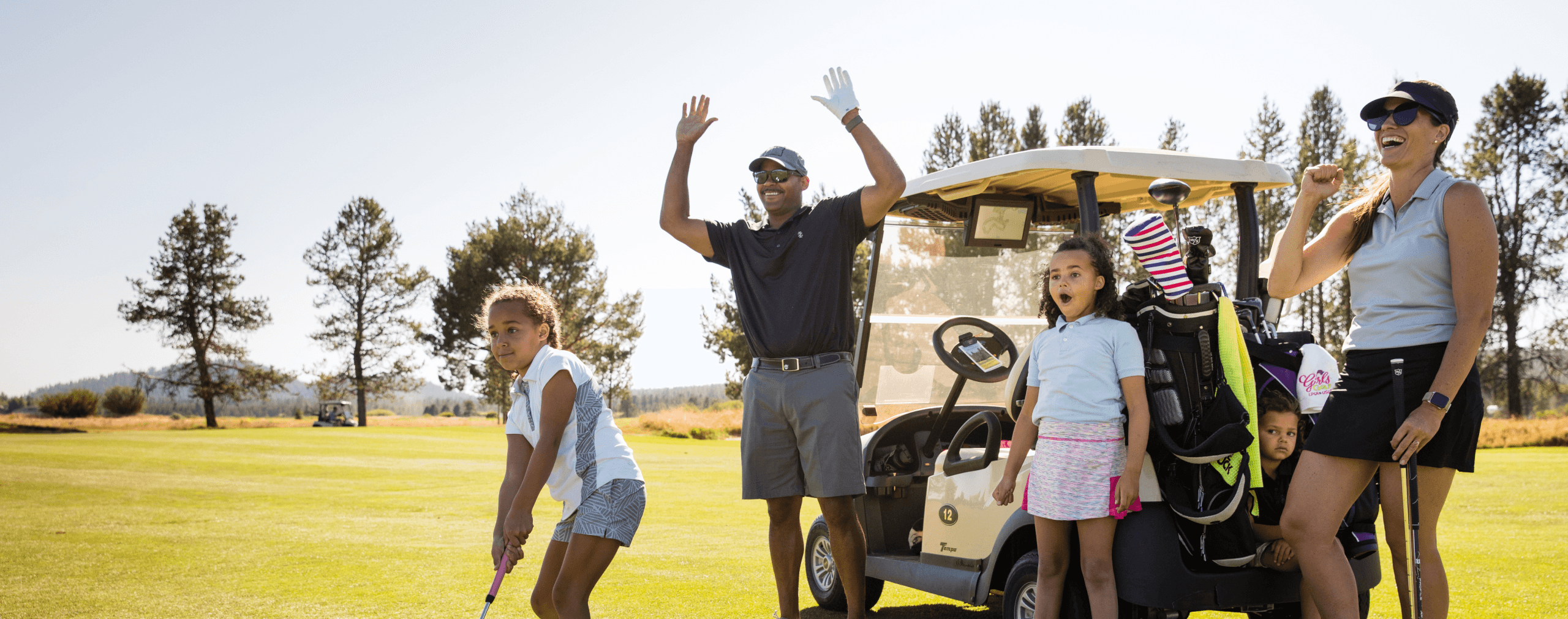 A family enjoys a day golfing; a man cheers with raised hands, two girls stand nearby, one preparing to swing, and a woman smiles next to a golf cart on a sunny course.