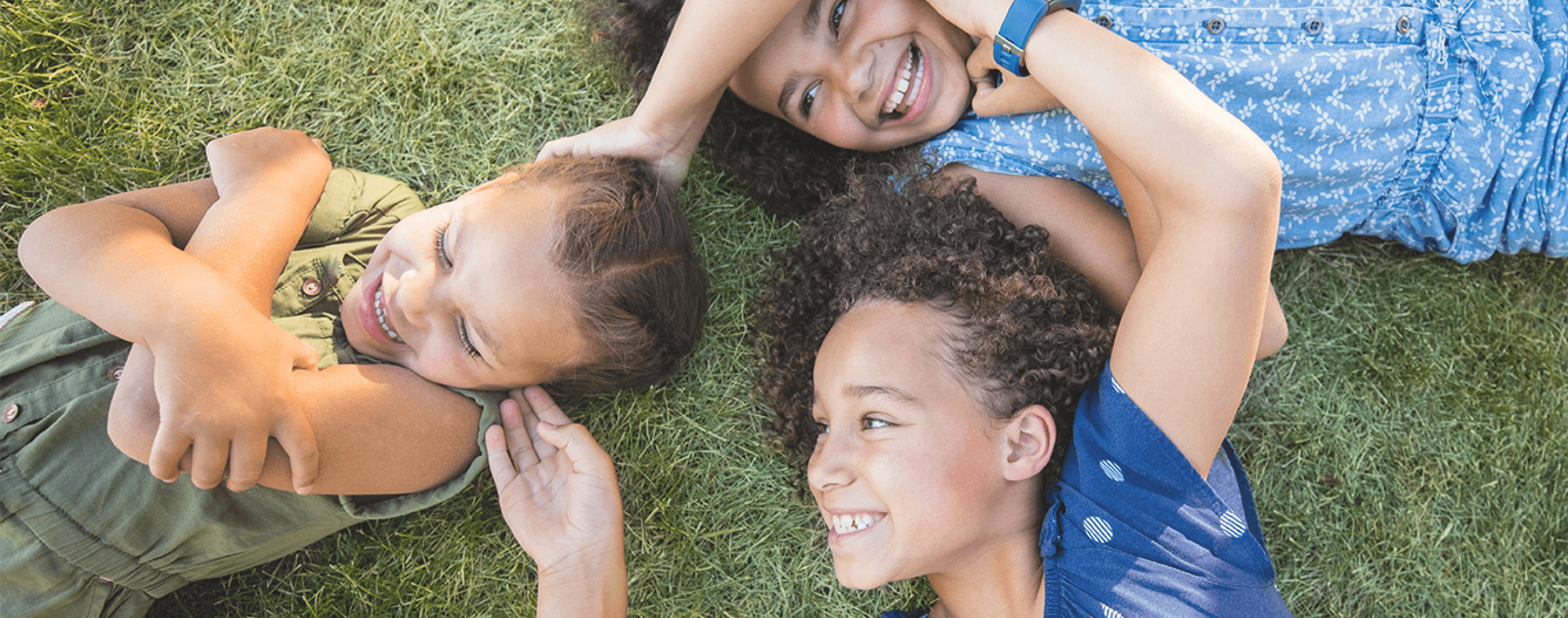 Three children lie on grass in a circle, smiling and laughing together. Their heads are close, and they appear happy and playful on a sunny day.
