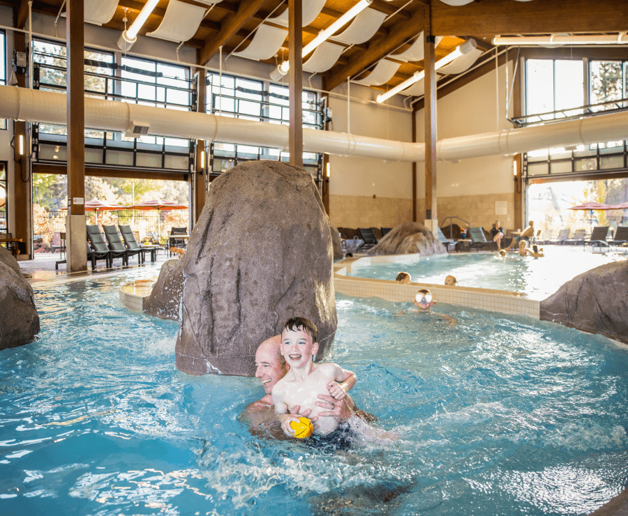 A man and young boy smile together in an indoor pool with rocks and lounge chairs around. Sunlight streams through large windows, and other people are swimming and relaxing in the background.