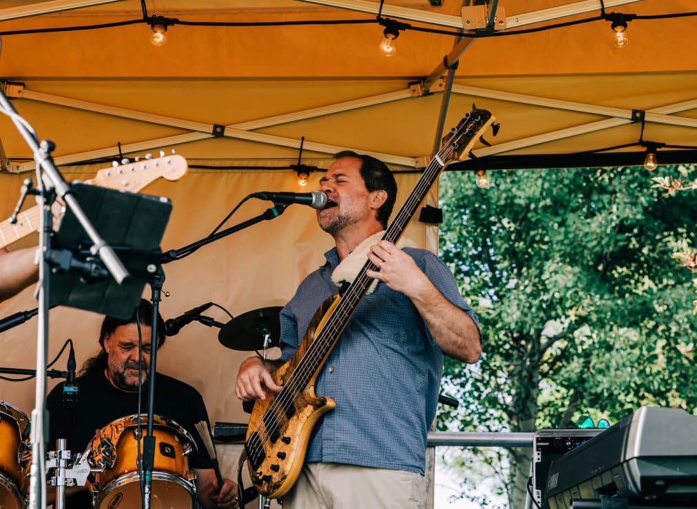 A man plays a bass guitar and sings into a microphone on an outdoor stage under a yellow canopy, with another musician on drums in the background.