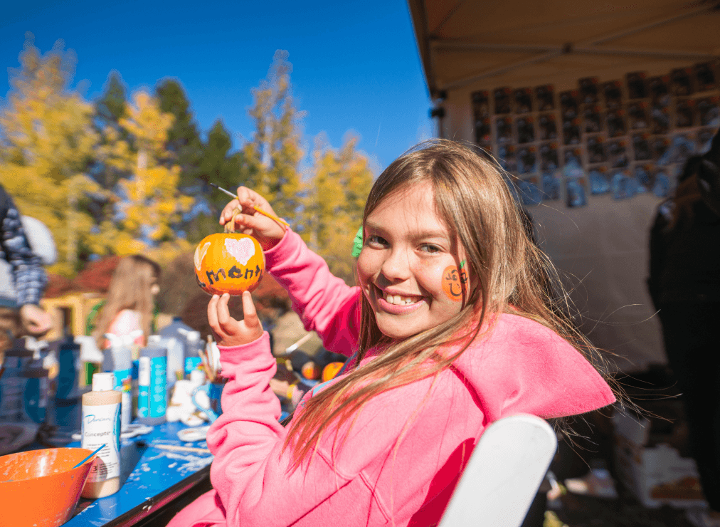A smiling girl in a pink hoodie paints a small pumpkin with mom and a heart. She sits outdoors at a table with paints, with colorful trees and people in the background.