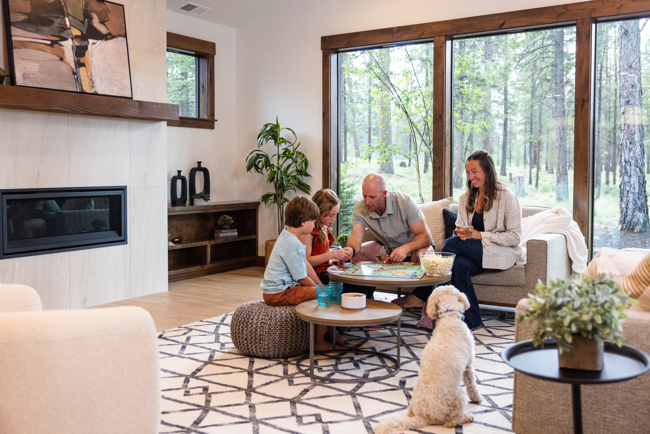 A family of four plays a board game in a cozy living room with large windows and a dog sitting on the rug, surrounded by trees outside.