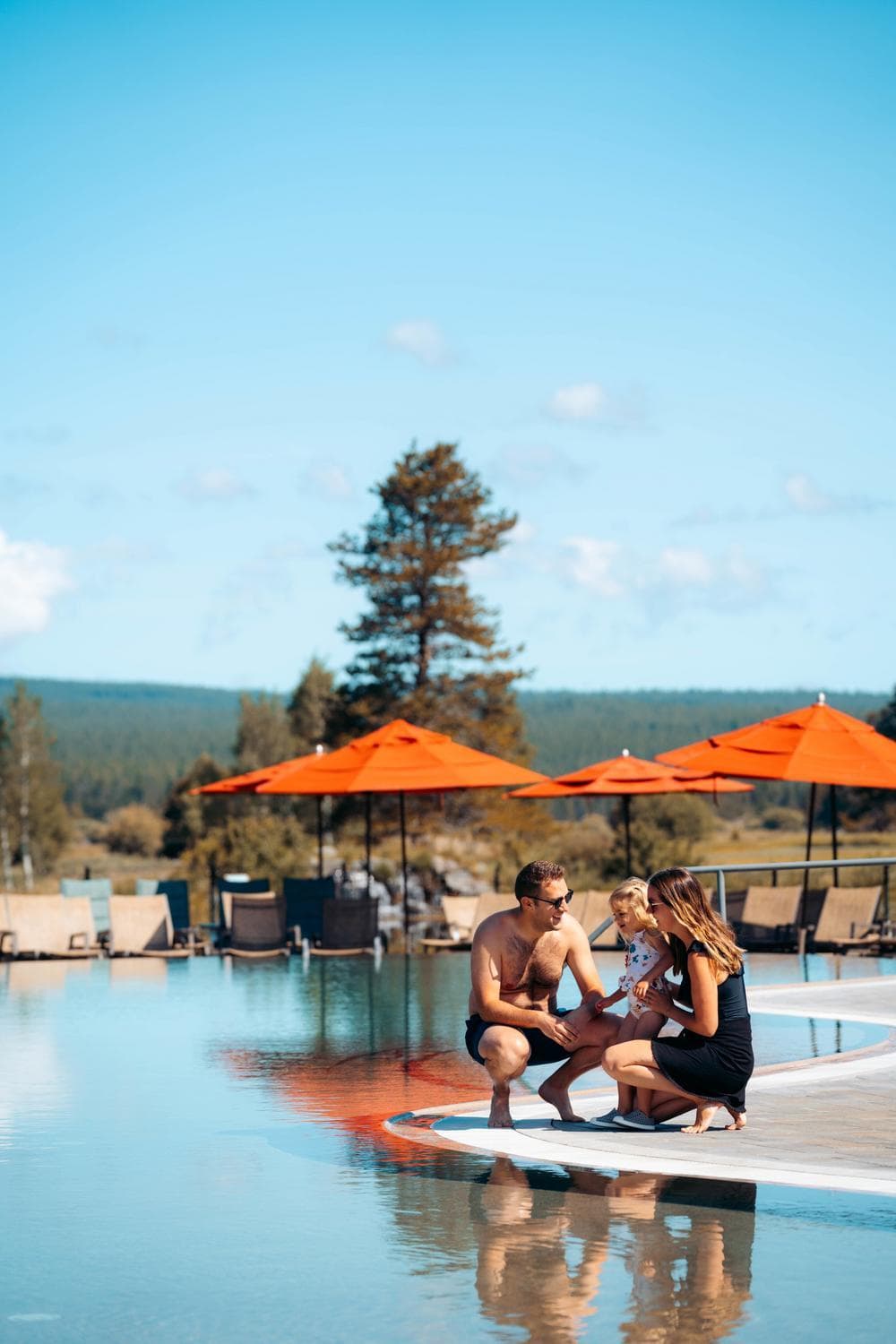 A family of three sits by a pool with orange umbrellas in the background. The adults and child smile and talk together. Trees and mountains are visible under a clear blue sky.