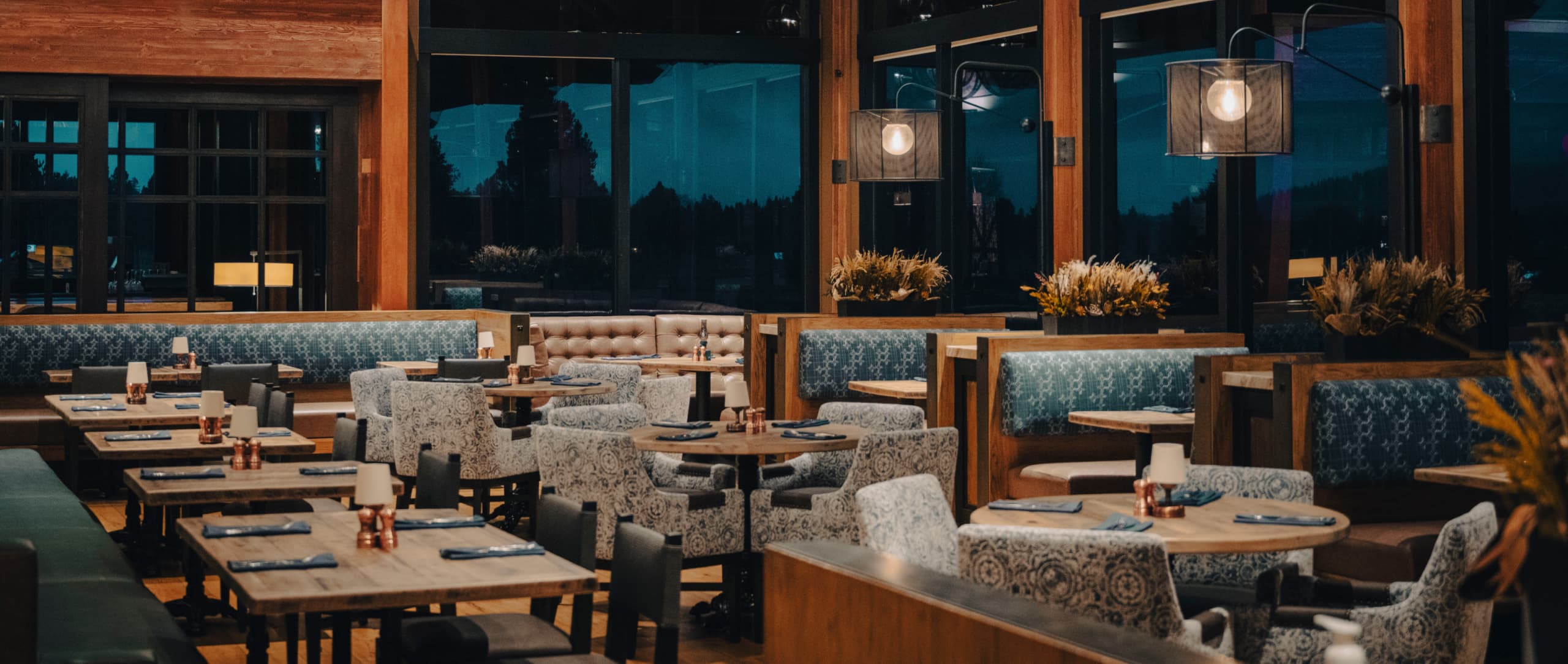 A modern restaurant interior with wooden tables, upholstered chairs, blue booths, large windows, and warm lighting, set for dinner with napkins and glassware on each table.