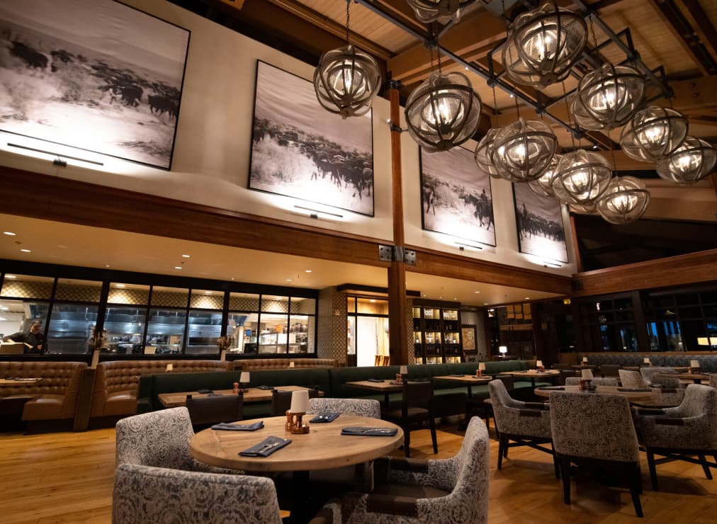 A modern restaurant with cushioned seating, round tables set with napkins, large black-and-white photos on wood-paneled walls, and decorative globe-shaped chandeliers hanging from the ceiling.
