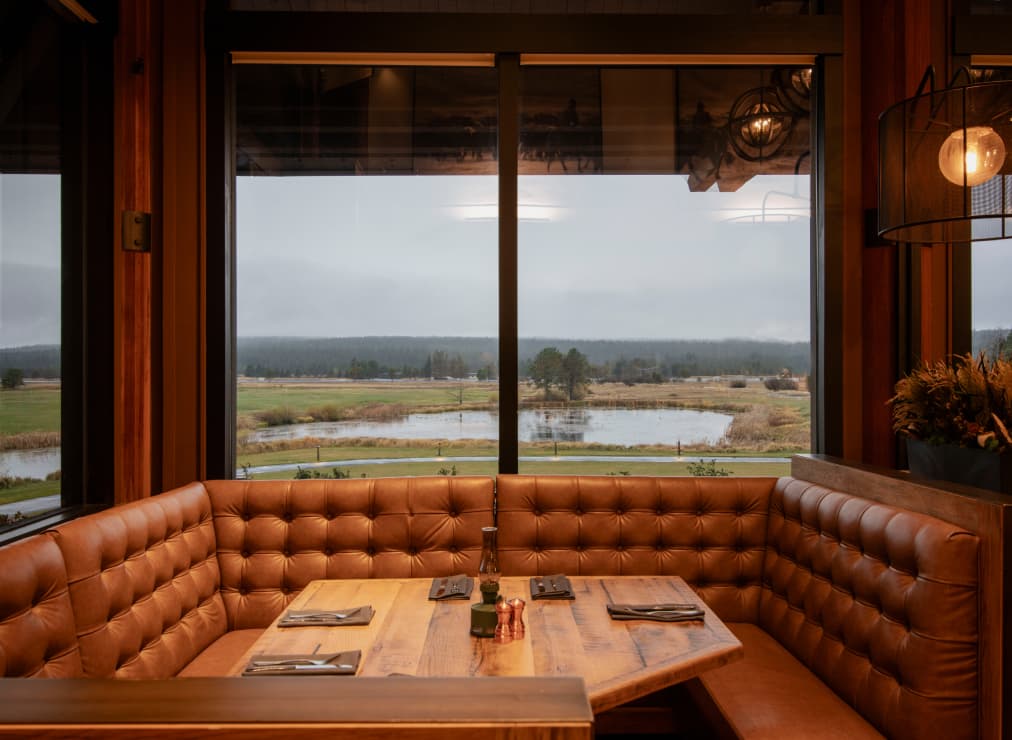A cozy restaurant booth with brown leather seating and a wooden table, set for four, overlooks a scenic pond and grassy landscape through large windows on a cloudy day.