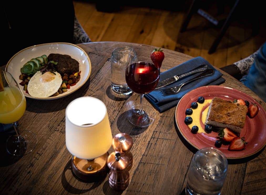 A wooden table set with a steak and fried egg dish, French toast with berries, glasses of red wine and juice, water, a lamp, and salt and pepper shakers.