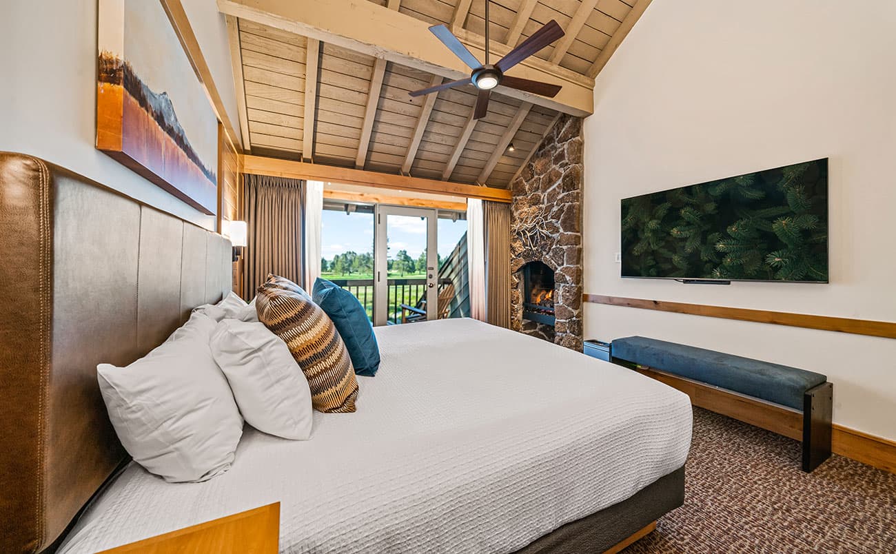 A cozy bedroom with a large bed, pillows, a wall-mounted TV, a stone fireplace, and a balcony with a scenic outdoor view. The room has high wooden ceilings and a bench below the TV.