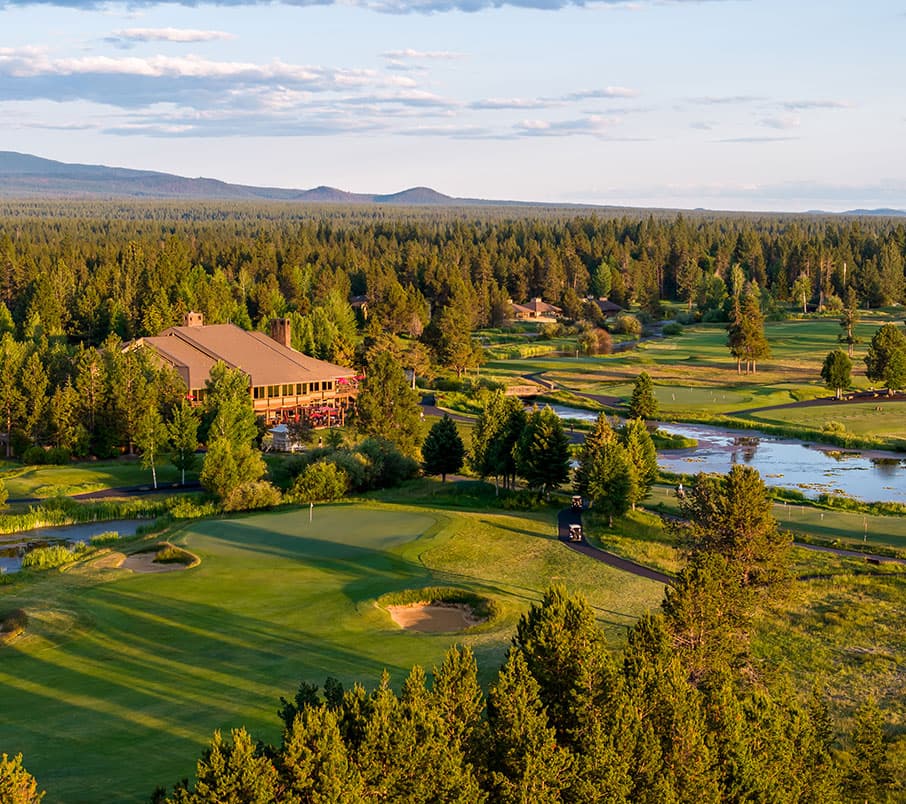 A golf course surrounded by trees and water features, with a clubhouse in the center and mountains in the distance under a partly cloudy sky.