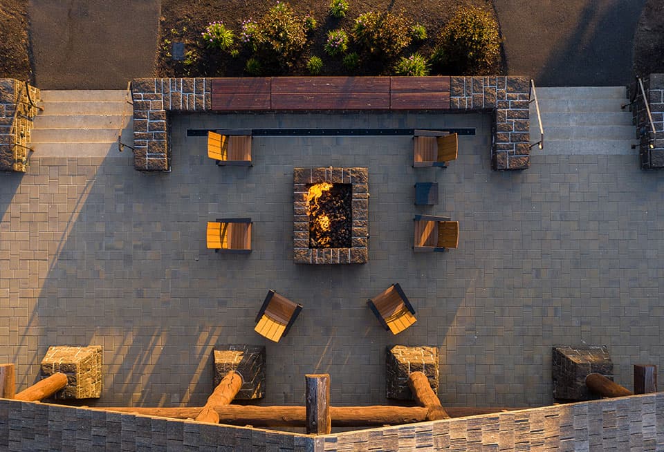 Aerial view of a stone patio with a square fire pit in the center, surrounded by wooden chairs and benches, bordered by landscaping and large timber posts.