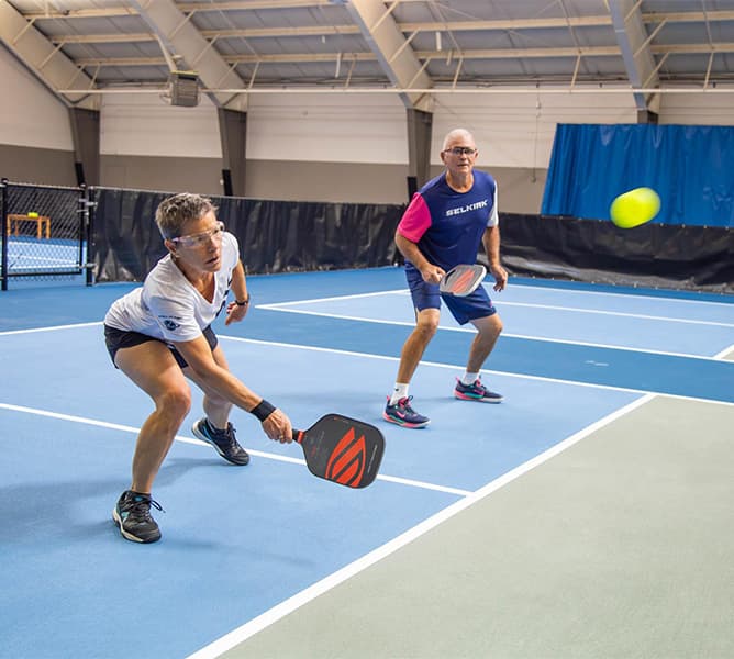 Two older adults play doubles pickleball on an indoor court. One lunges to hit a low ball with a paddle while the other stands ready behind, both wearing sportswear and glasses.