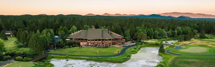 Large lodge-style building surrounded by trees, with a pond and golf course in the foreground and mountains in the background at sunset.