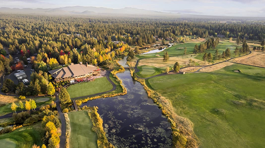 Aerial view of a golf course surrounded by trees, a clubhouse, ponds, and mountains in the background under a cloudy sky.