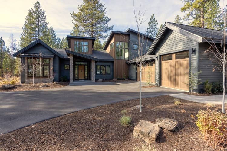 Modern two-story house with dark wood siding, large windows, and a spacious driveway surrounded by trees and landscaping. The sky is partly cloudy and the yard is mostly bare with some small plants.