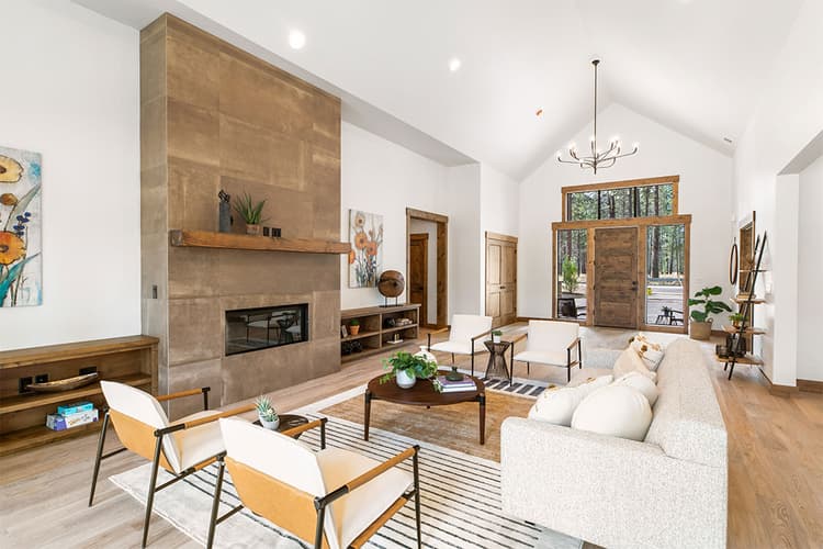 Modern living room with high ceilings, tan fireplace, large windows, neutral furniture, light wood flooring, and minimal decor. Light fills the space, highlighting plants, artwork, and cozy seating areas.