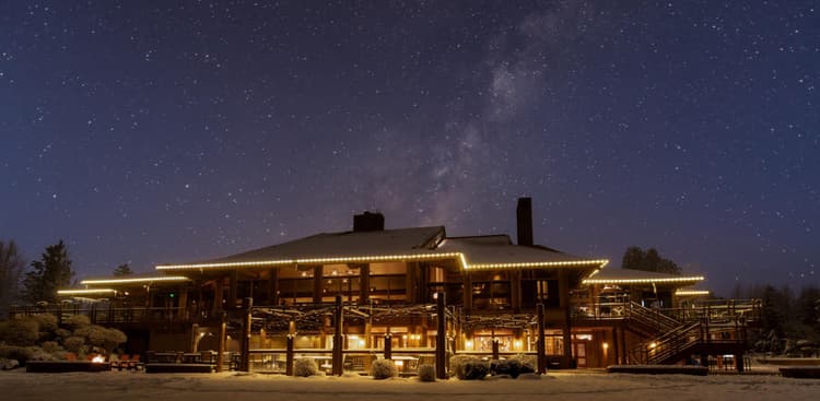 A large, warmly lit lodge with snow outside sits under a clear night sky filled with stars and the Milky Way. Trees surround the building, and yellow lights outline the roof.